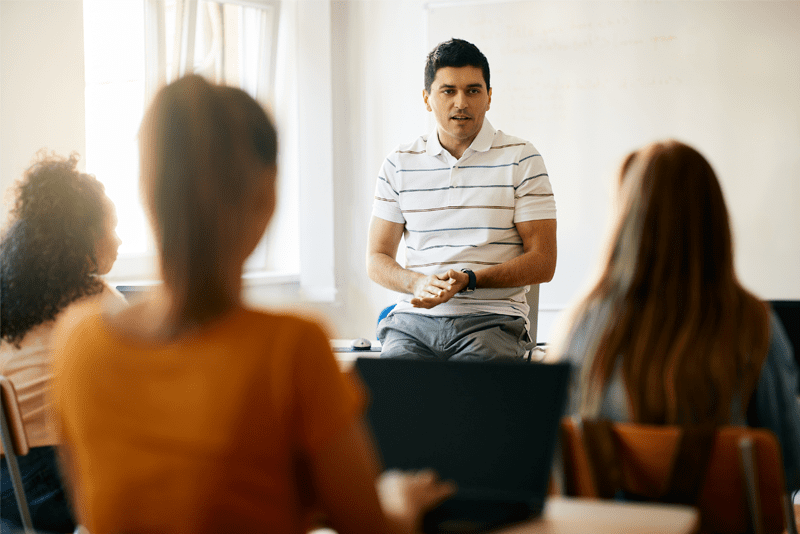 Teacher sitting on a desk in a classroom addressing three students