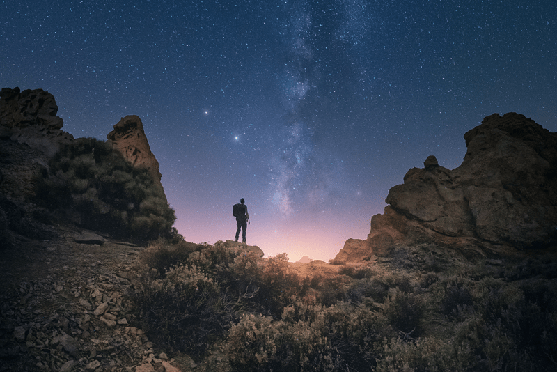 Man with backpack surrounded by mountains staring at a pink-purple sky